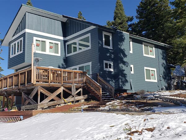 Blue-gray mountain home with vertical siding and wraparound deck in snow